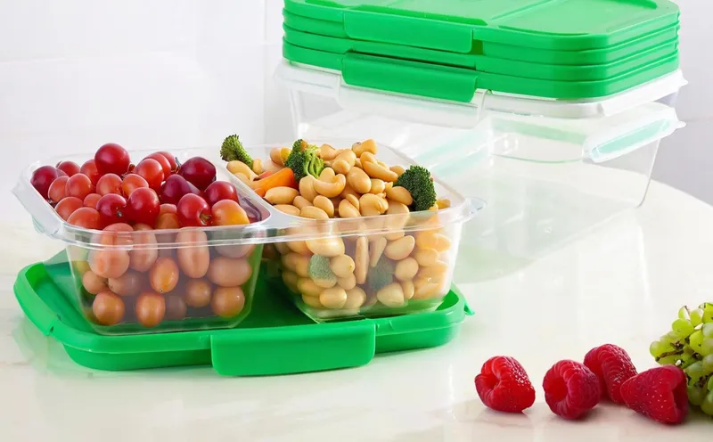 Plastic containers filled with tomatoes and beans, with green lids and additional stacked containers in the background, alongside fruits on a table.