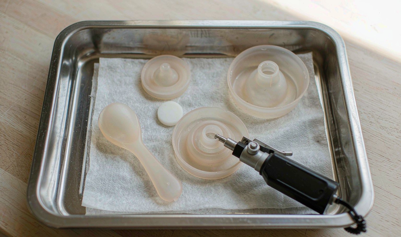 Pale translucent silicone baby products on a stainless tray with a durometer gauge pressed against one teat