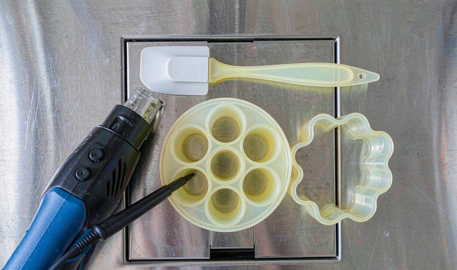 Pale grey silicone kitchenware on a stainless bench, a flexible spatula, a six-cup muffin mould and a trivet, with a hot-air gun and thermocouple probe